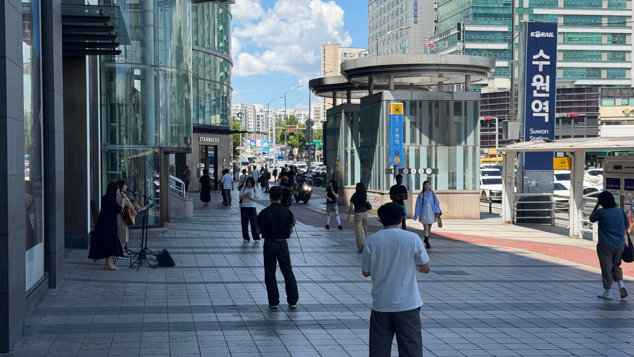 공연자 두 명과 보행자, 수원역 표지가 함께 보이는 보도 전경